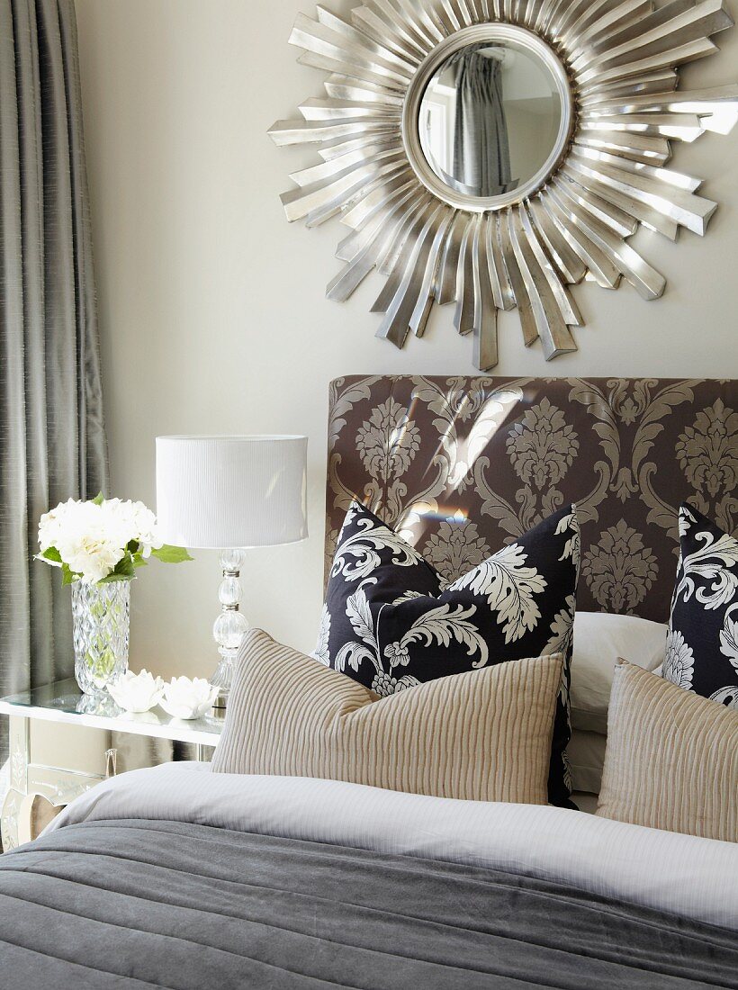 Kissen auf Bett vor gepolstertem Kopfteil mit Ornament Muster, darüber Spiegel mit strahlenförmigem Rahmen