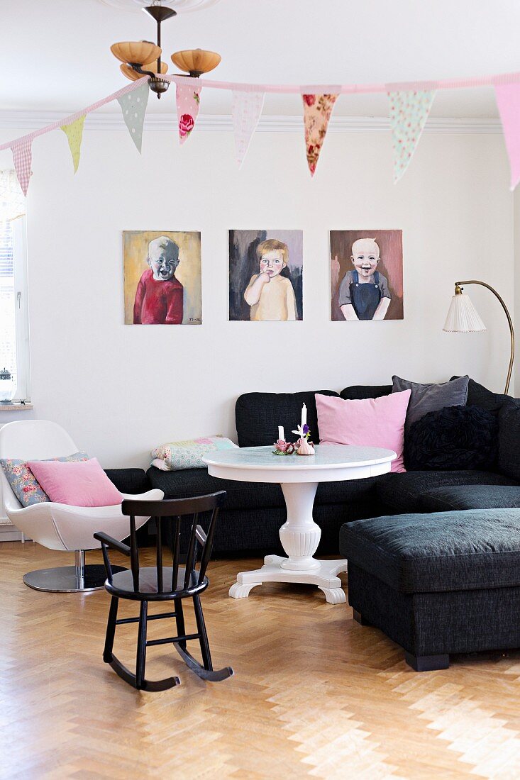 Wohnzimmerecke mit gemalten Kinderportaits über schwarzem Polstersofa, weisser runder Tisch und Kinderschaukelstuhl auf gepflegtem Eichenparkett