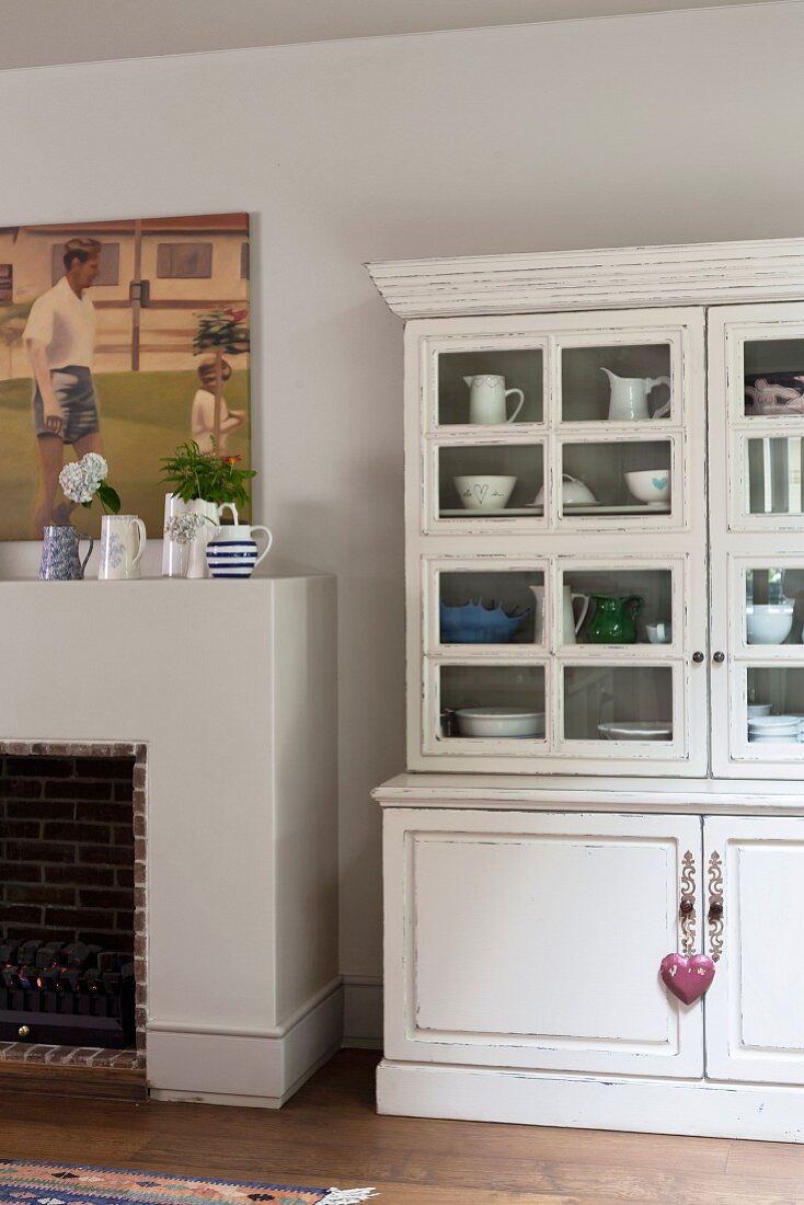 White, vintage dresser with glass-fronted top section next to open fireplace