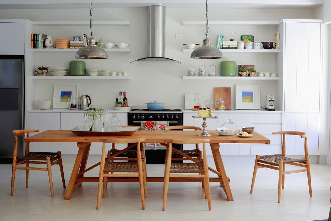 Solid wood dining table and chairs in open-plan kitchen with white fitted cabinets and open-fronted shelving