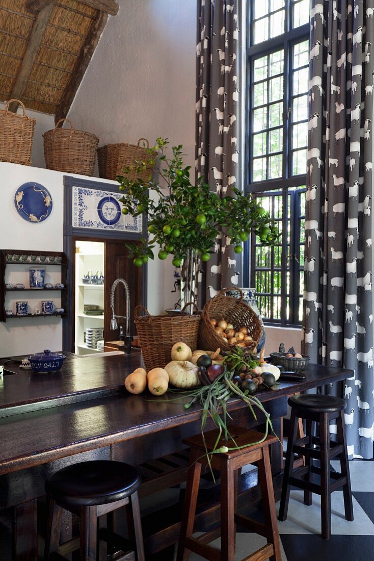 Vegetables and baskets on dark wood kitchen counter, bar stools and tall lattice window with floor-length curtains