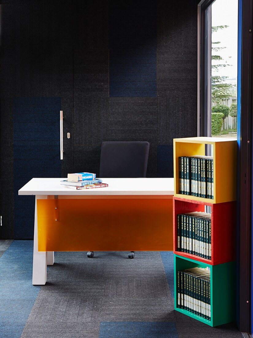 Bunte Holzmodule mit Büchern übereinander gestapelt, dahinter Schreibtisch mit orangefarbener Kunststoffplatten