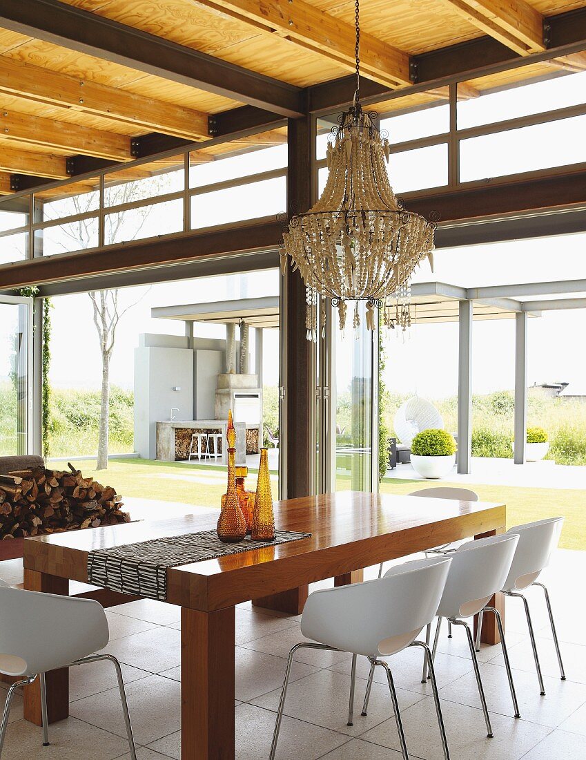 Chandelier above dining set with elegant wooden table and shell chairs; view into garden through steel and glass facade