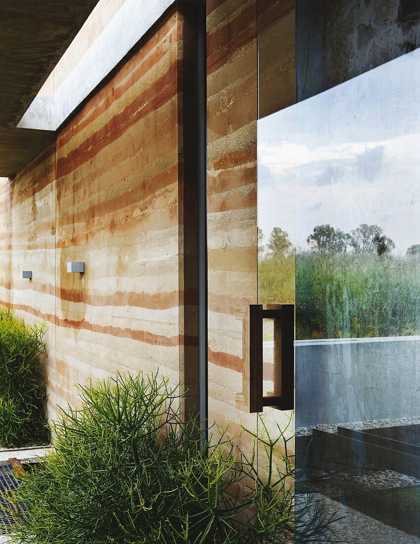 Facade of modern house with rammed-earth structure and reflection of surrounding countryside in glass door