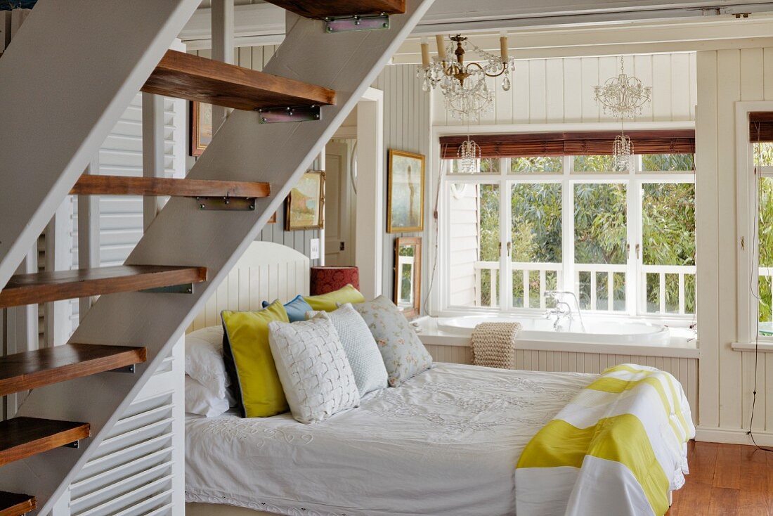 Schlafzimmer mit Doppelbett neben eingebauter Badewanne vor Fenster in holzverkleidetem Schlafzimmer, im Vordergrund Holztreppe