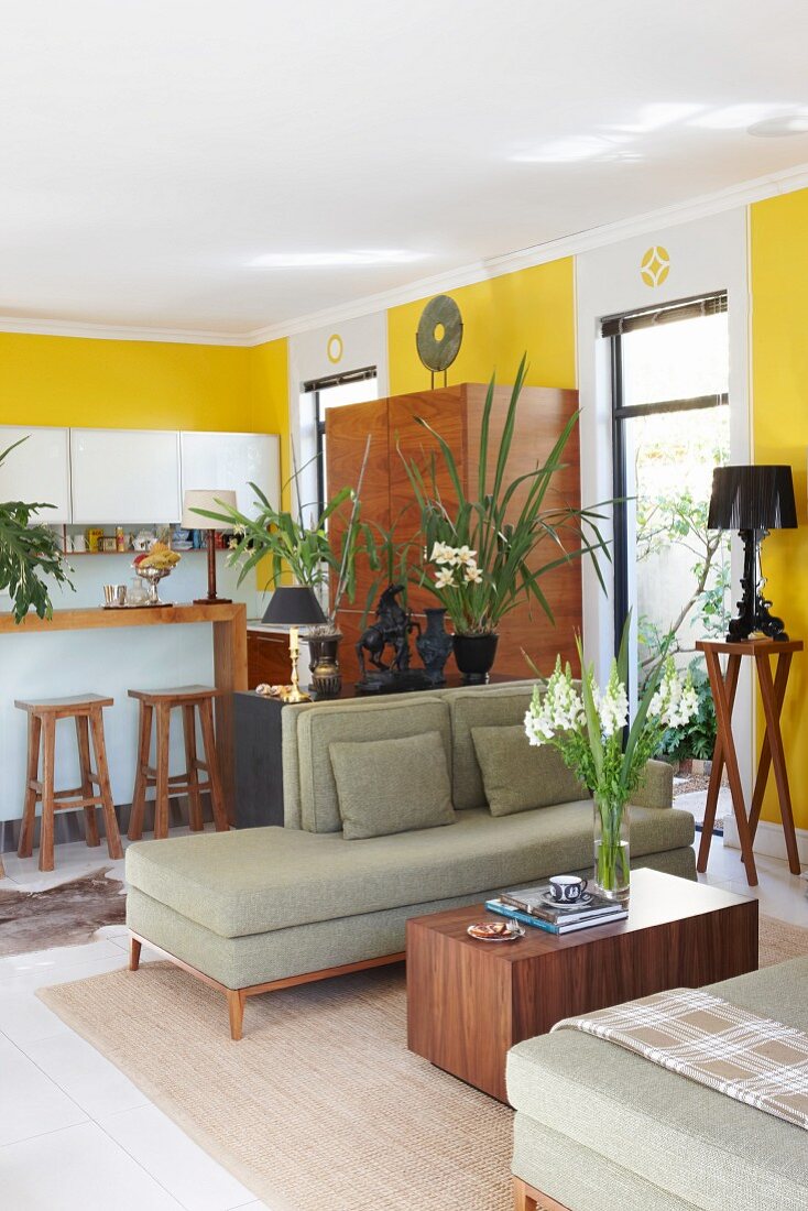 Cubic wooden coffee table between retro chaise longue and kitchen area in open-plan interior with yellow-painted walls