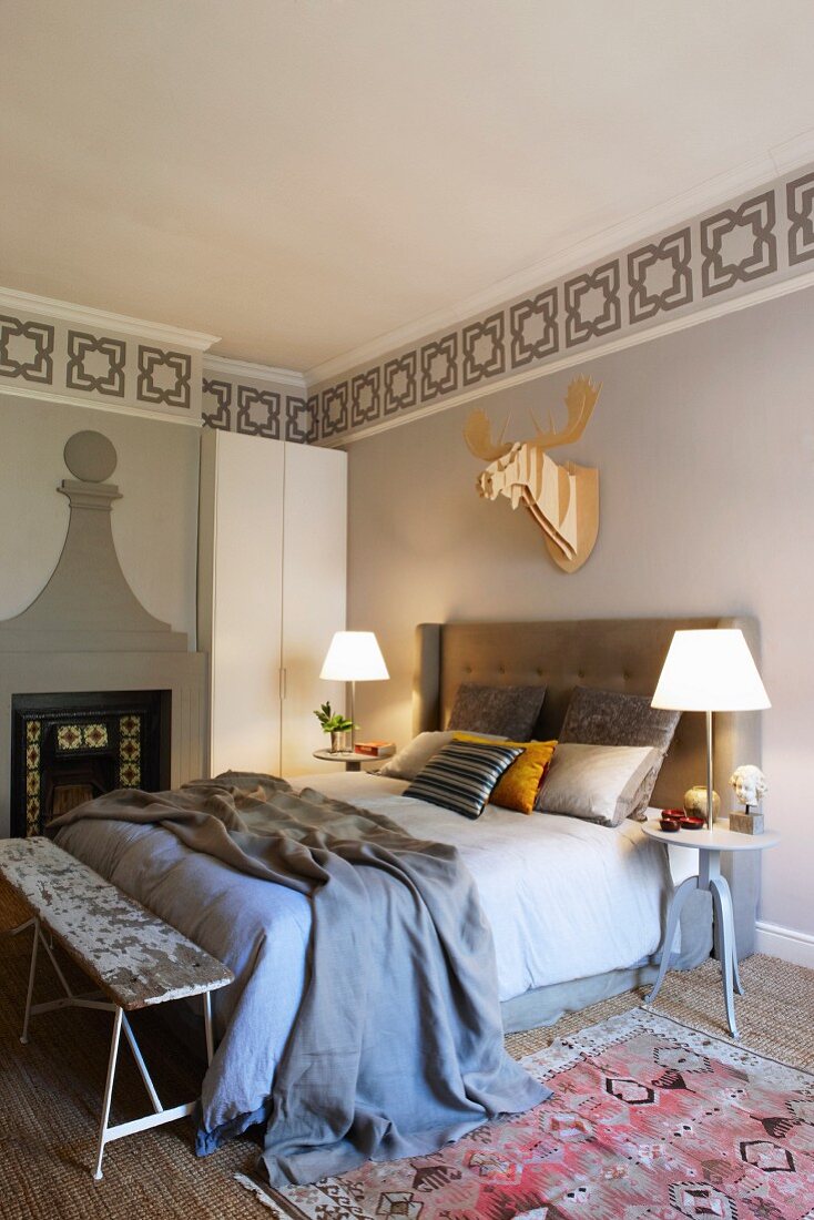 Doppelbett mit gepolstertem Kopfteil, seitlich Nachttisch mit Tischleuchten, an Wand Bordüre in Schablonentechnik, darunter stilisierte Tiertrophäe