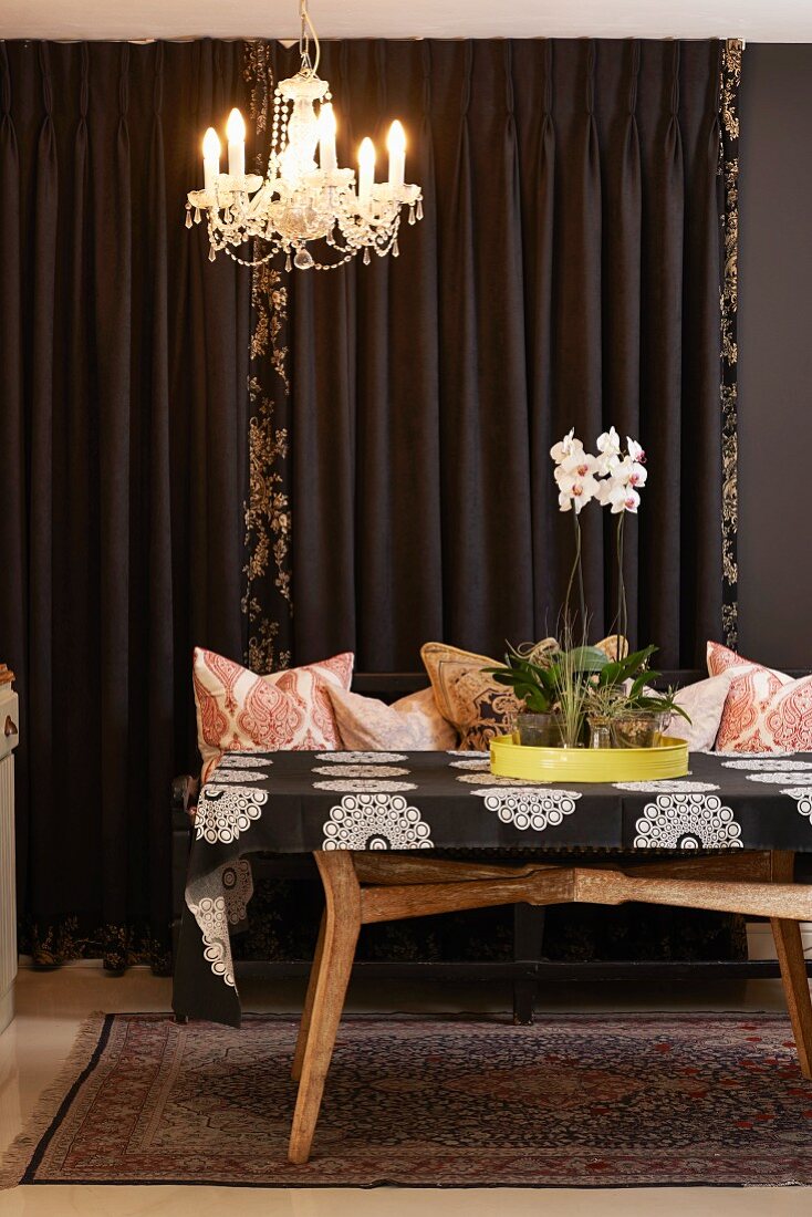 Retro Holztisch mit Orchideen auf gelbem Tablett, vor braunem Vorhang, an Decke Kronleuchter mit Perlenschmuck