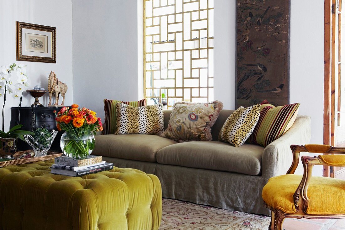 Gepolsterter Couchtisch mit gelbgrünem Bezug und bunter Blumenstrauss in Vase gegenüber Sofa am Fenster mit gitterartigem Holzgestell, an der Seite antiker gepolsterter Stuhl