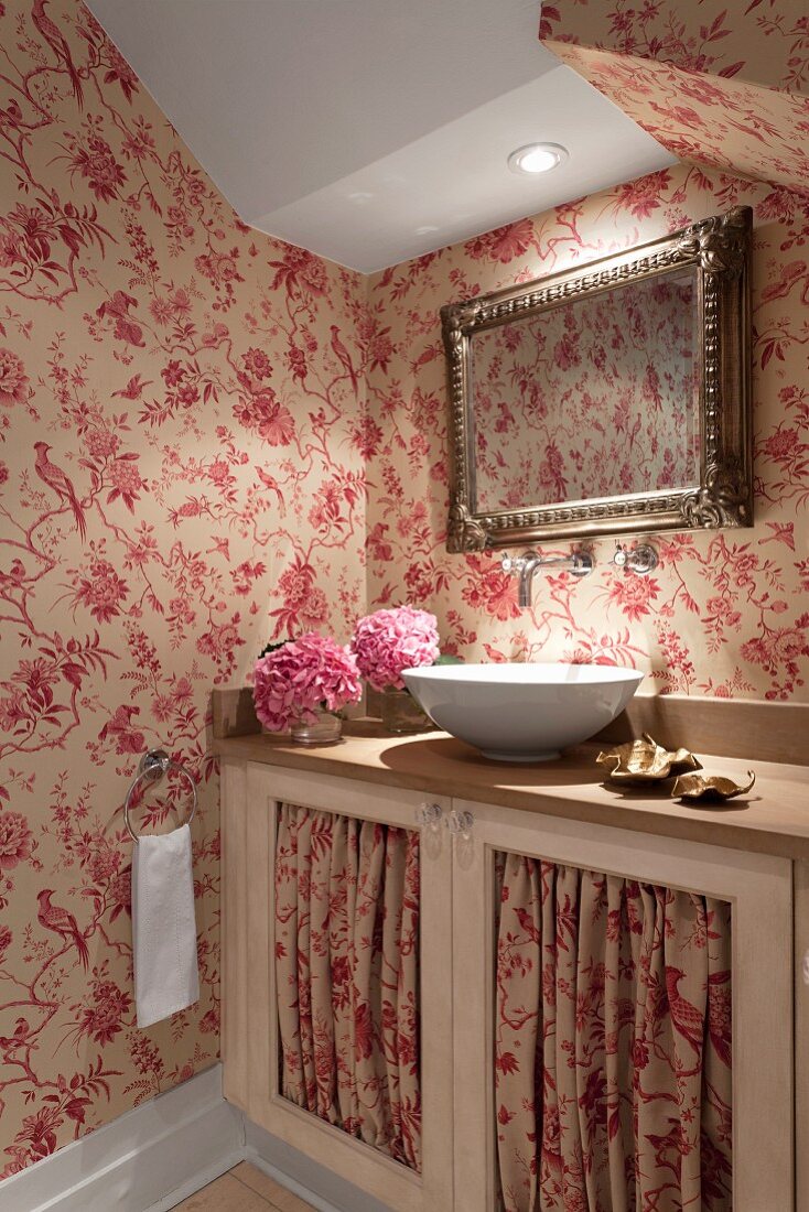 Illuminated washbasin on base unit with red and white patterned fabric panels in doors and matching wallpaper in attic room