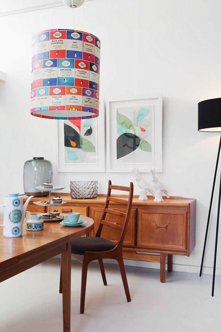 Chair at set table below pendant lamp with colourful lampshade; sideboard below framed pictures on wall in background