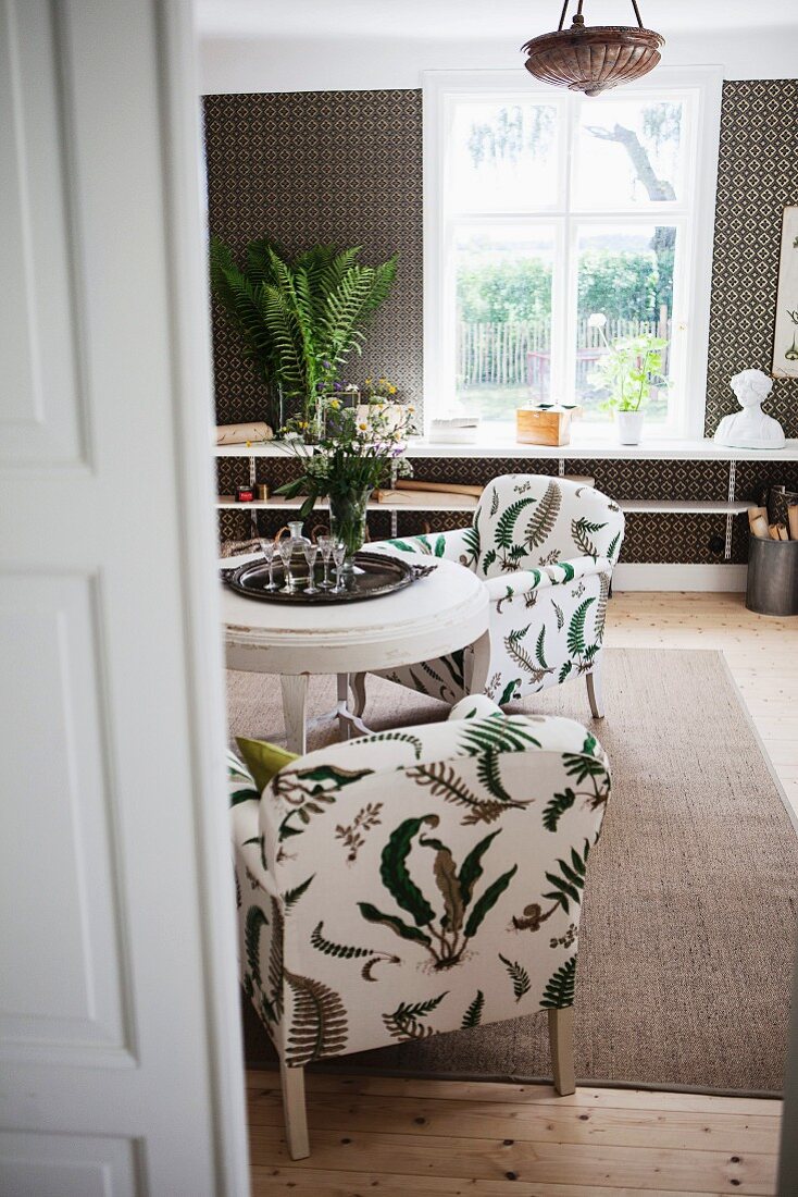 View through open door of armchairs with pattern of stylised leaves on upholstery in interior with rustic ambiance