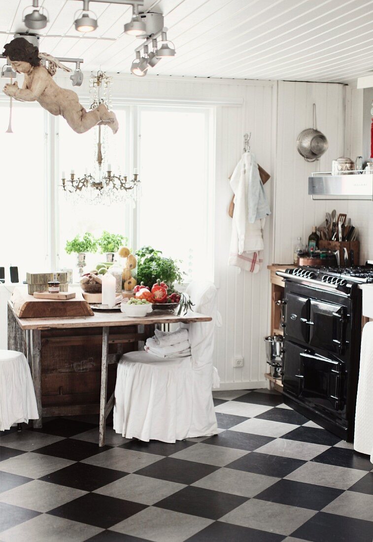Rustic kitchen with chequered floor, chairs with loose white covers at dining table in front of window and vintage cooker to one side