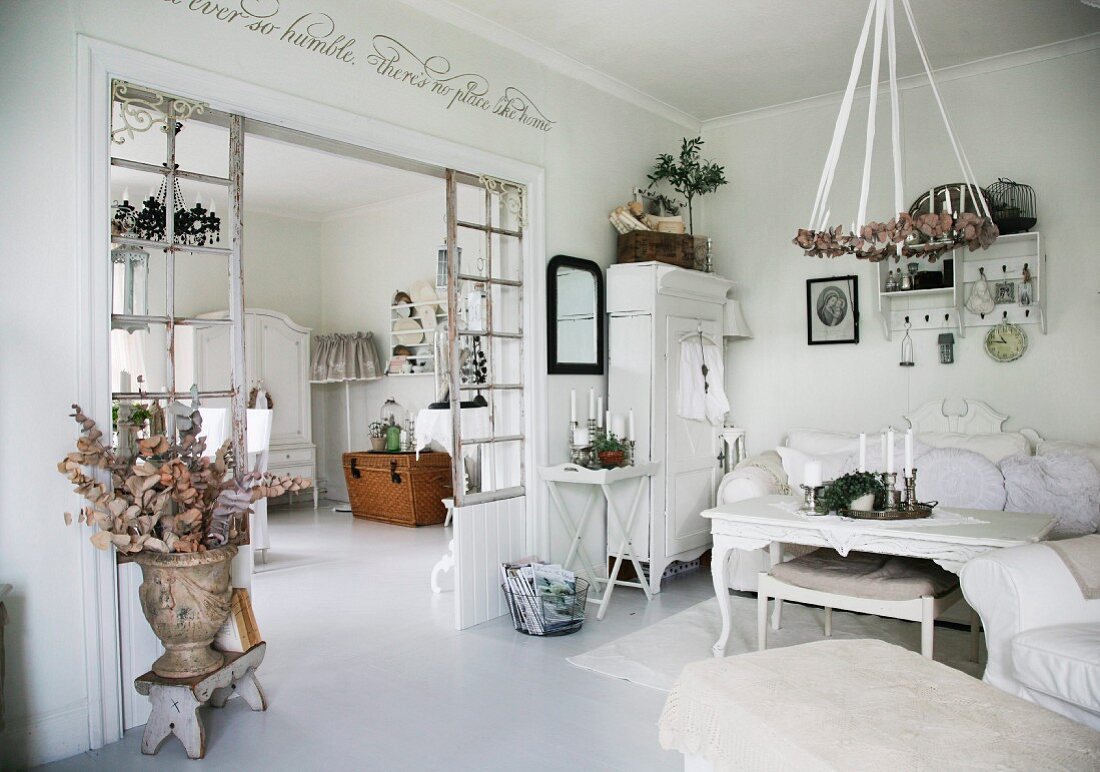 Ländlicher Wohnraum in Weiß, an Decke aufgehängter Kranz über Couchtisch, seitlich offener Durchgang mit Sprossen-Raumteiler und Blick ins Esszimmer