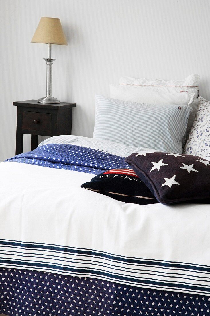 Bett mit blau weiss gemusterter Tagesdecke und Kissenstapel, seitlich Nachttisch mit Leuchte