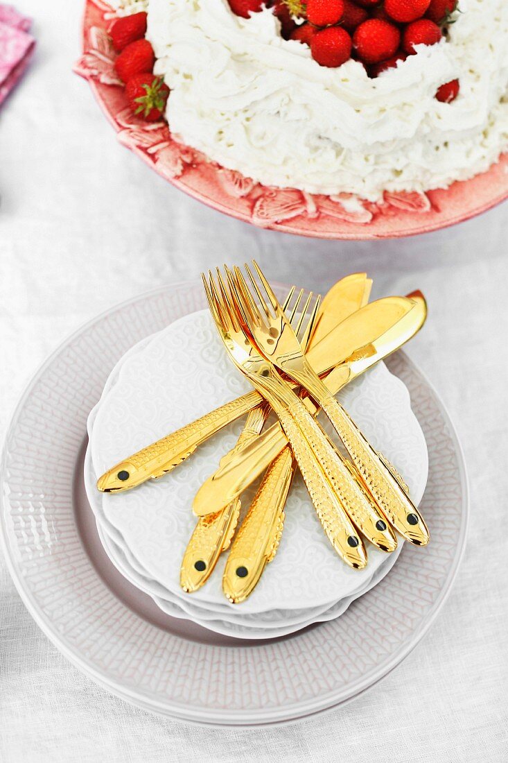 Goldfarbenes Dessertbesteck mit Fischmotiv auf Tellerstapel neben Sahne-Erdbeerkuchen