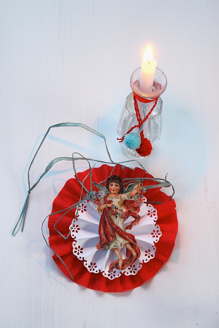 Weihnachtlich geschmückte Glasvase als Kerzenständer vor Papierschmuck mit Engel