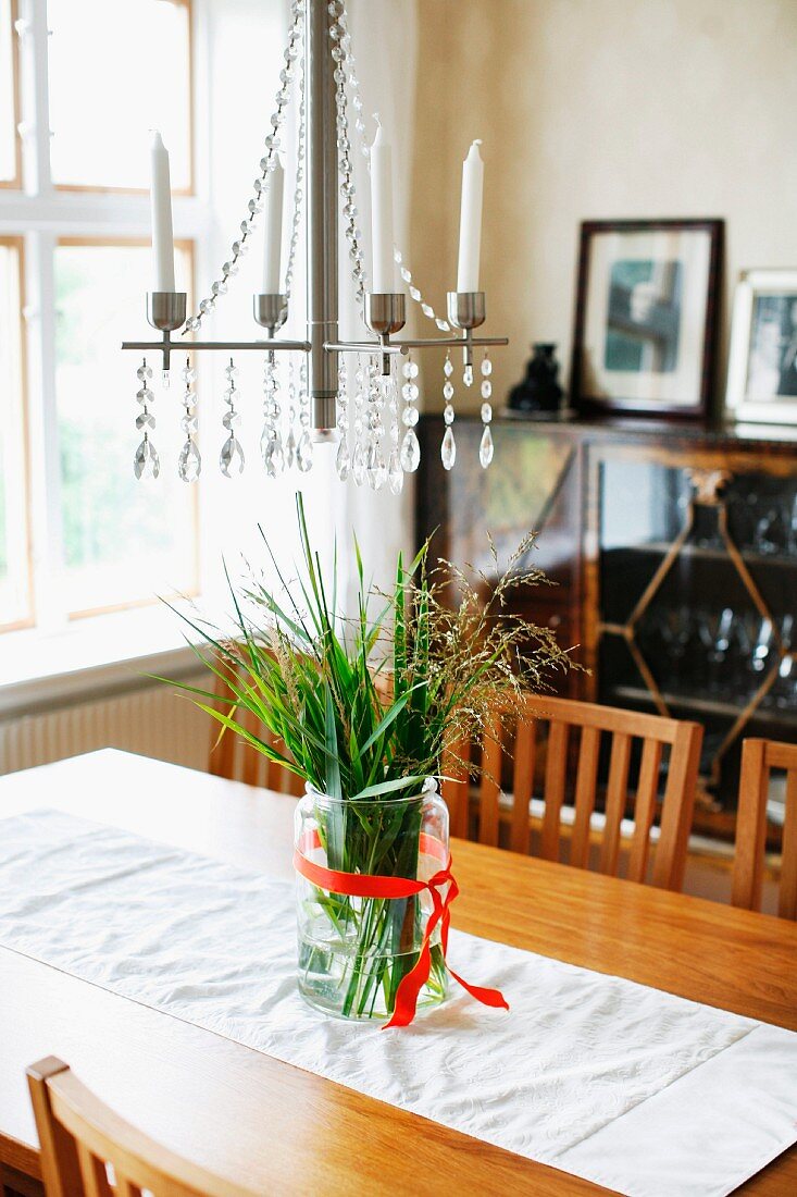 Candle chandelier with white candles above glass vase of ornamental grasses and white runner on wooden dining table