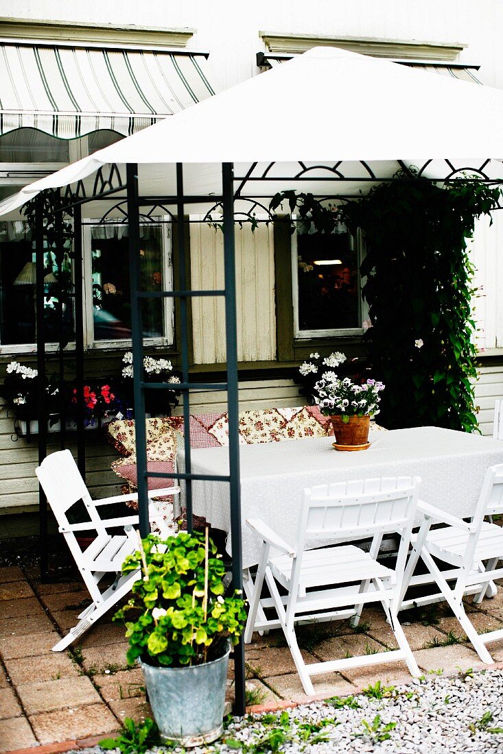 Terrassenplatz vor Wohnhaus, weiss lackierte Holzstühle um Tisch unter Pergola mit Zeltdach aus weißem Stoff