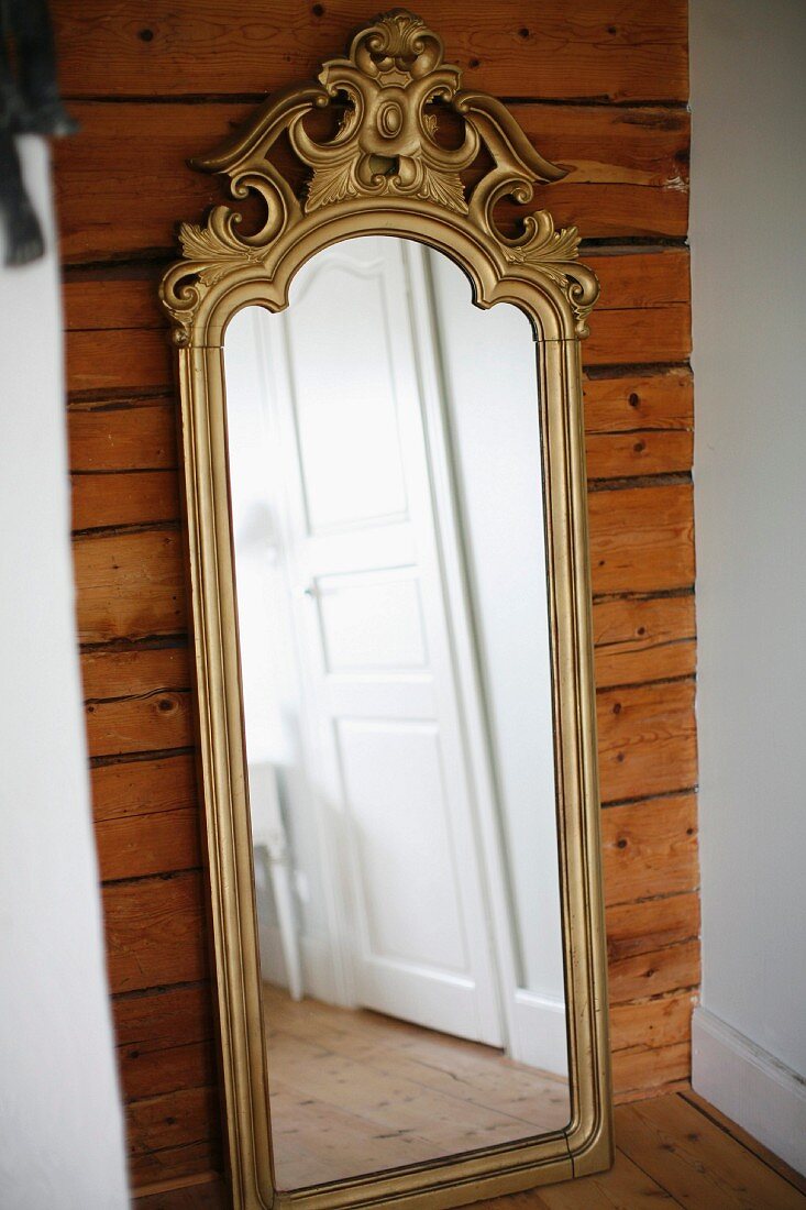 Antique, full.length mirror with gilt, carved frame leaning against wooden wall