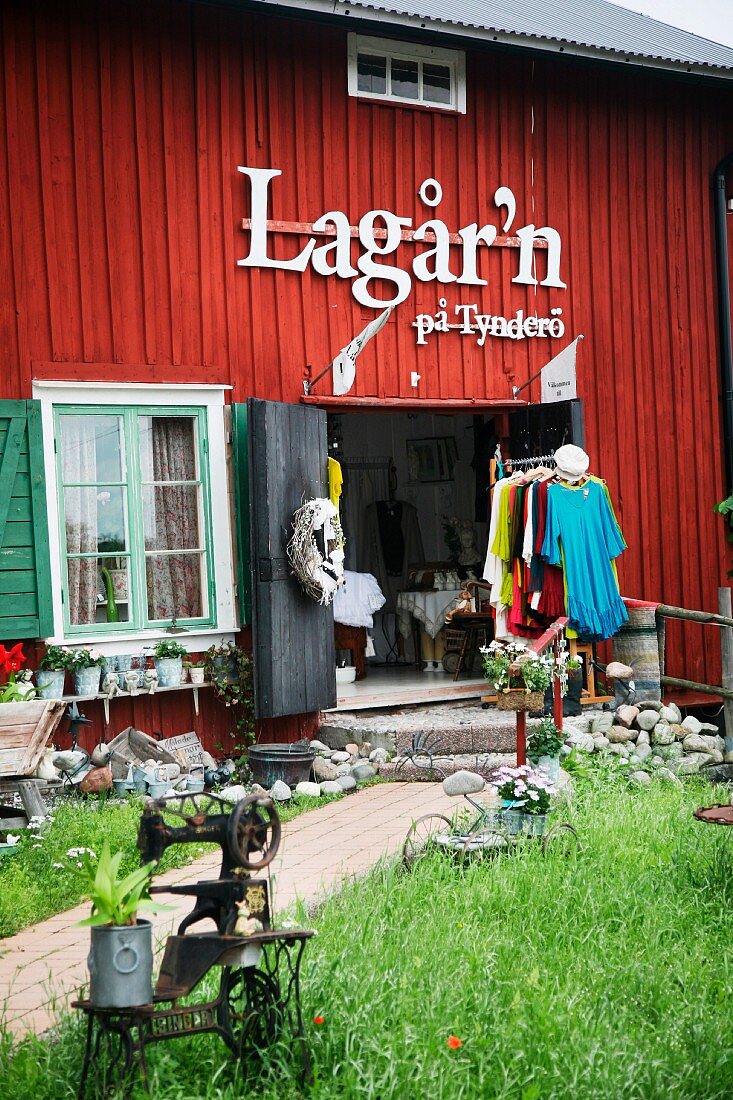 Offener Kleiderladen in rotem, rustikalem Holzhaus, davor Kleiderständer, im Vordergrund Vintage Nähmaschine in der Wiese