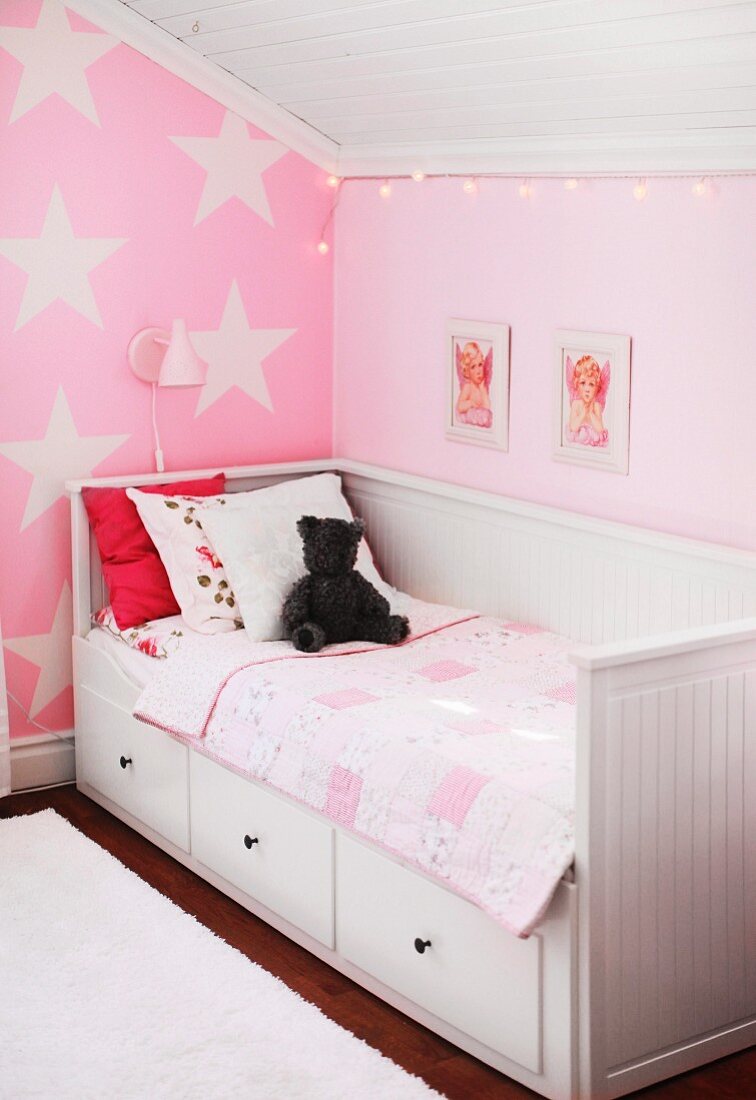 White wooden bed with drawers in pink, child's attic bedroom with star-patterned wall and pictures of cherubs