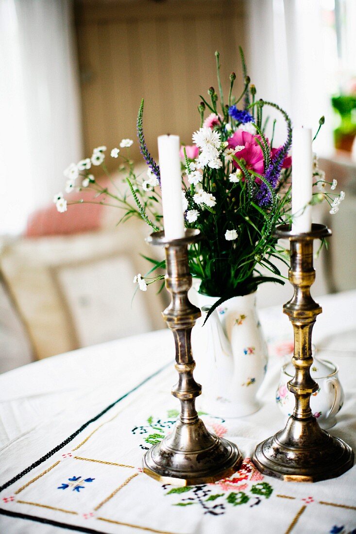 White candles in brass candlesticks and vase of garden flowers on table with tablecloth