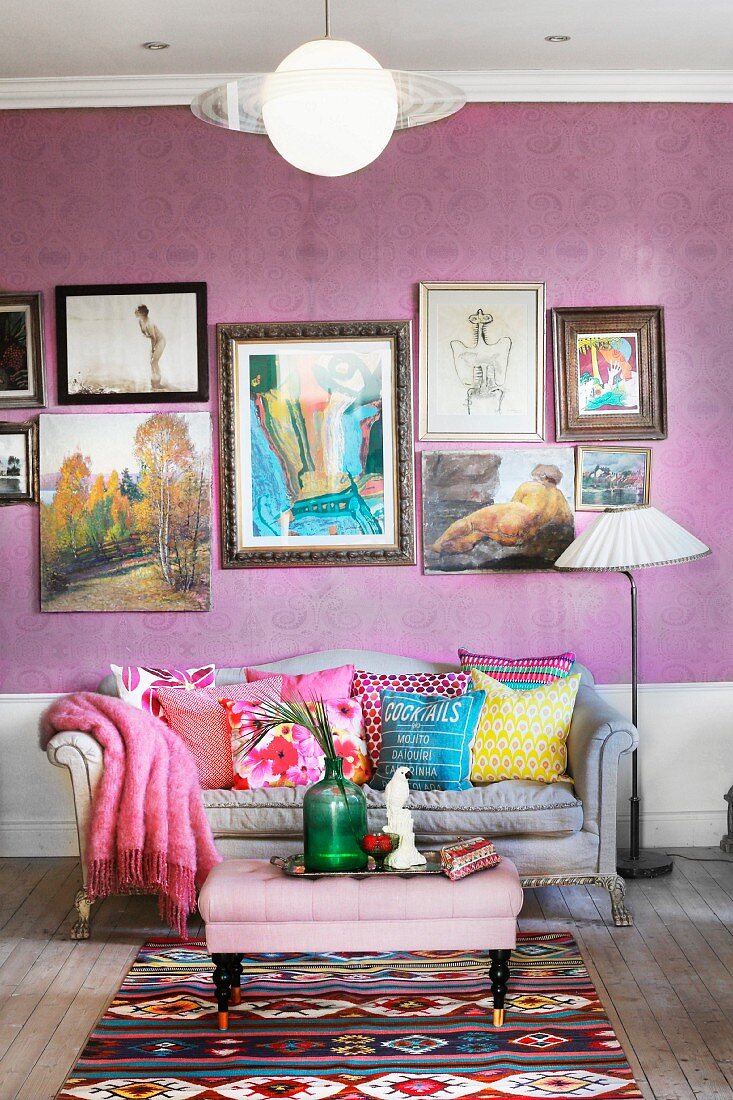 Polstertisch und geschwungene Couch mit bunten Kissen vor lila Tapete an Wand mit gerahmter Bildersammlung