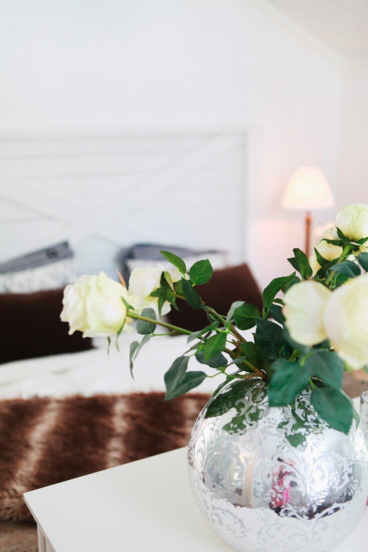 weiße Rosen in bauchiger, verzierter Bauernsilbervase; im Hintergrund Doppelbett