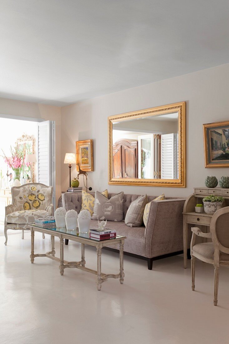 Elegant interior in pale shades; antique coffee table, arranged scatter cushions on sofa and gilt-framed mirror on wall