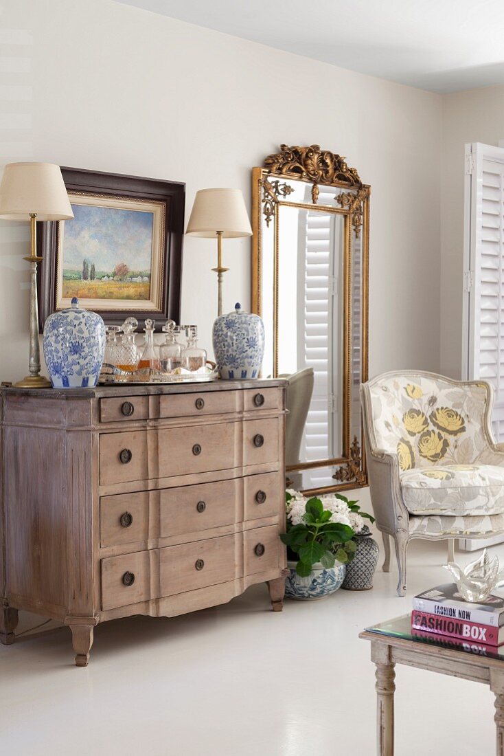 Table lamp with pale lampshade and lidded jars on wooden chest of drawers next to armchair and full-length mirror