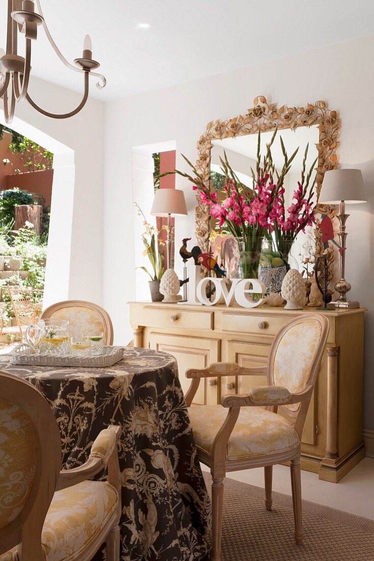Eleganter Essplatz mit Stühlen im Rokoko Stil, im Hintergrund Gladiolenstrauss und Tischleuchten auf Kommode aus hellem Holz
