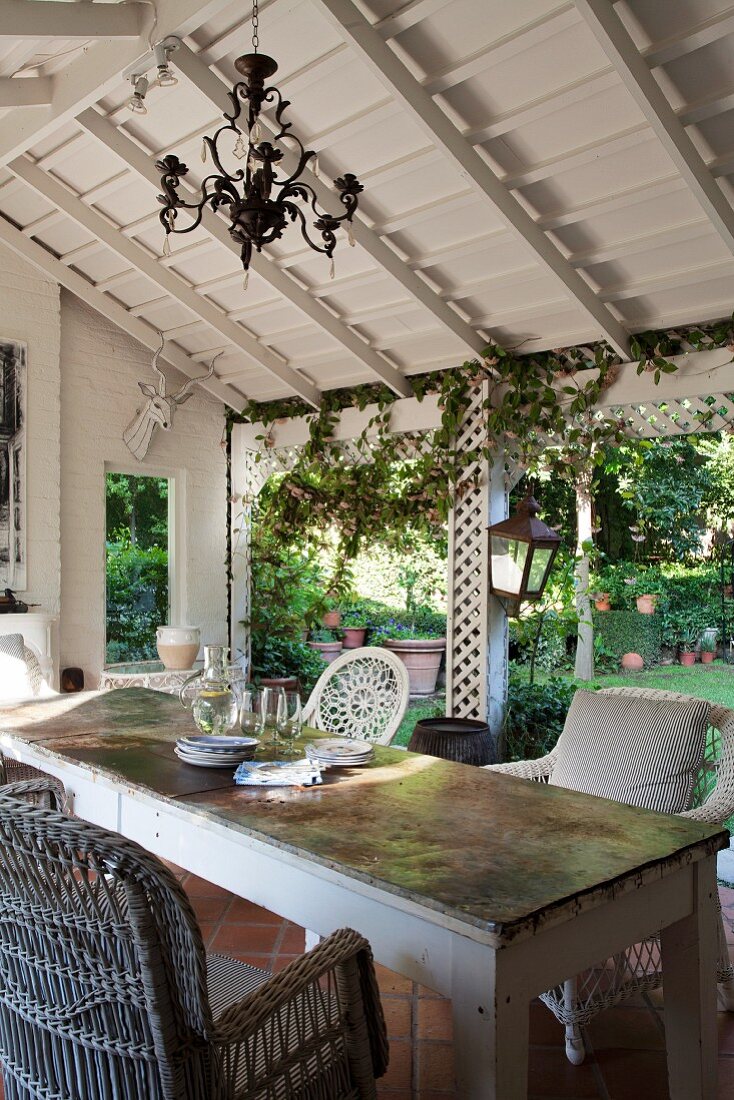 Kronleuchter, Korbstühle und Tisch mit Patina auf überdachter Landhaus-Terrasse; Blick in Garten unter berankten weißen Holzgittern hindurch