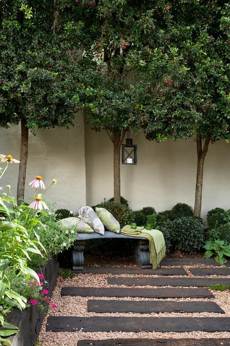 Courtyard with dark wooden sleepers set in gravel floor; trees against wall in background
