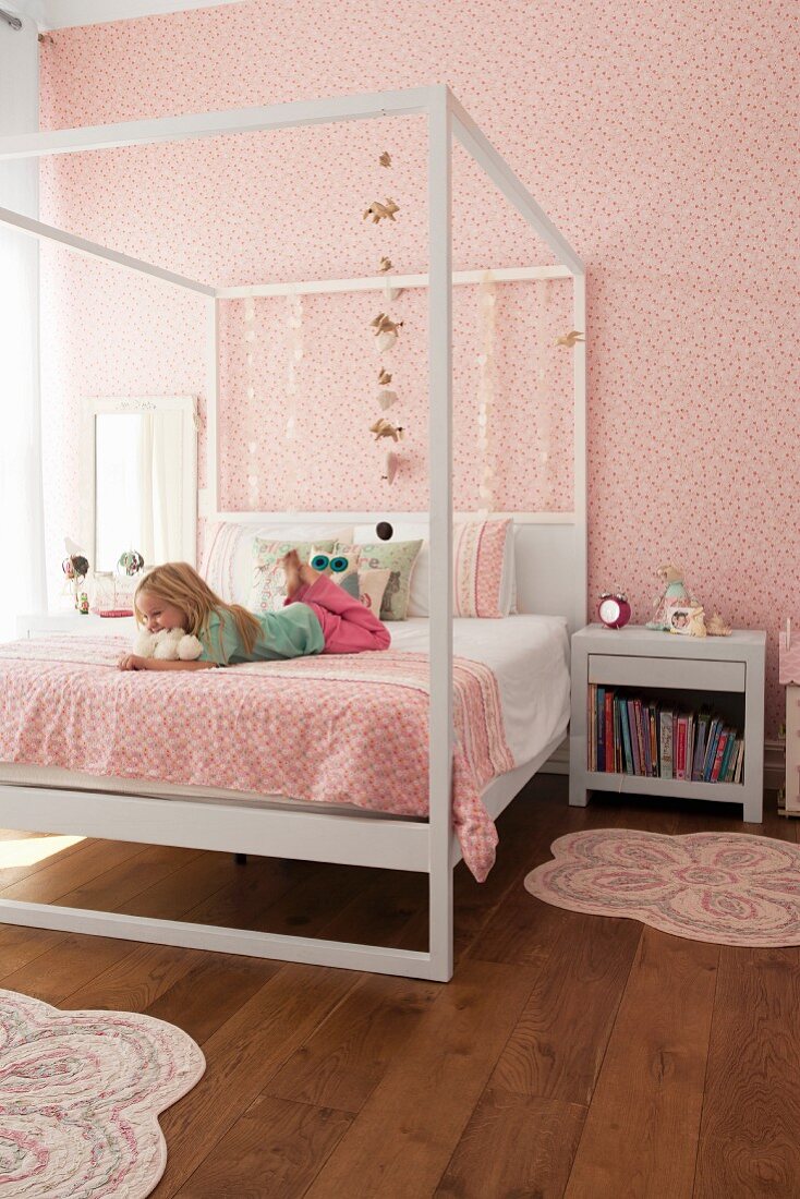 Girl lying on bed with white four-poster frame; bedspread and wallpaper with matching patterns