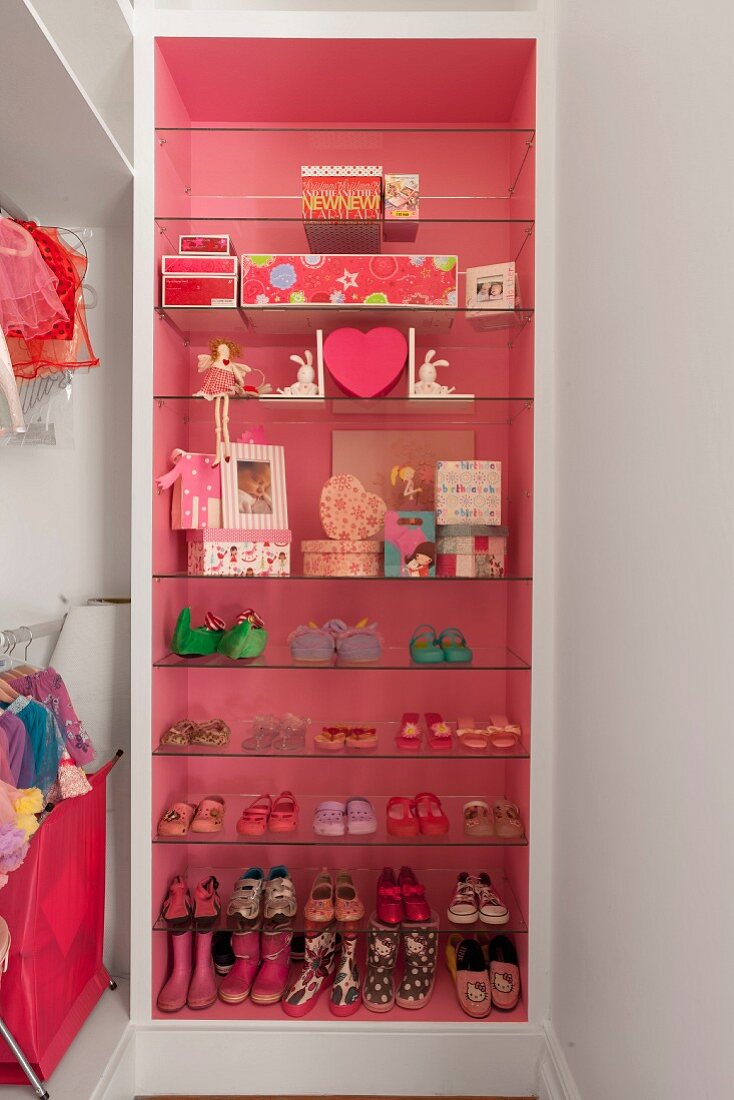 Offener Regalschrank mit Mädchenschuhen und dekorative Aufbewahrungsboxen auf Glasböden, Hintergrund pinkfarben