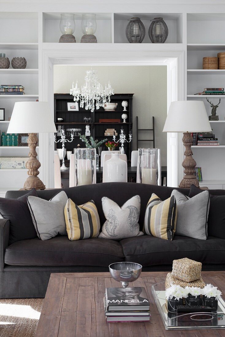 Coffee table, black sofa with arranged scatter cushions, two table lamps and white fitted shelves surrounding doorway in background