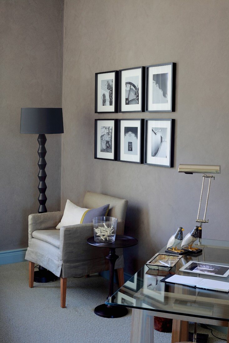 Teilweise sichtbarer Schreibtisch, gegenüber Sessel und Stehleuchte mit schwarzem Schirm, vor braun getönter Wand mit Fotos