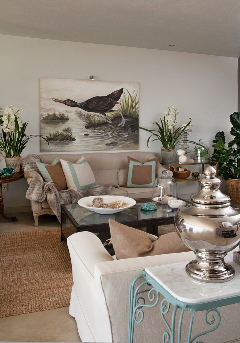 Silver vessel on side table against sofa backrest and picture of aquatic bird on wall in living room with country-house ambiance