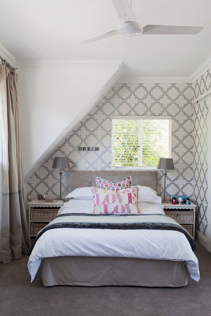 Double bed below sloping ceiling in bedroom with classic, white and grey patterned wallpaper