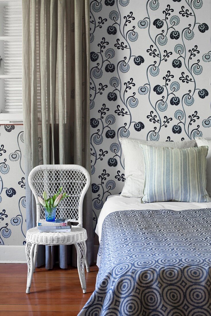Weisser Geflechtstuhl neben Bett mit gemusterter Tagesdecke und Tapete an Wand mit weiss-blauem, stilisiertem Blumenmuster im Schlafzimmer