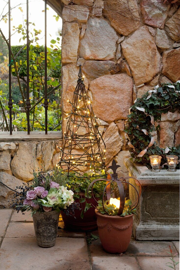 Christmas atmosphere in garden, lit candle in flower pot and obelisk covered in fairy lights in front of stone wall