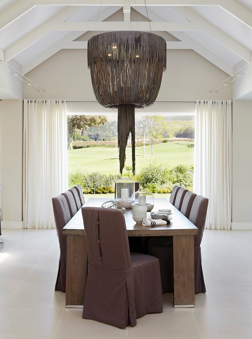 Chairs with purple loose covers in dining area below pendant lamp with leather lampshade and view through French windows