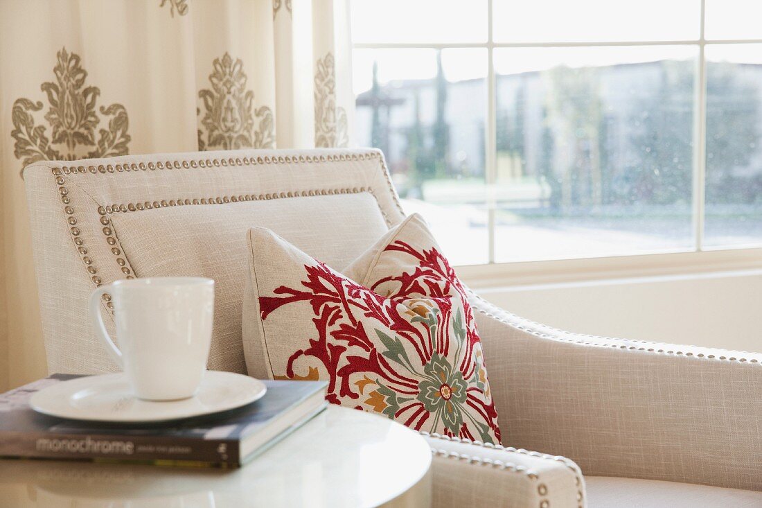 Teetasse auf Buch und Beistelltisch neben Sessel mit Ziernägeln auf Bezug und gemustertes Kissen neben Fenster mit Stadtblick