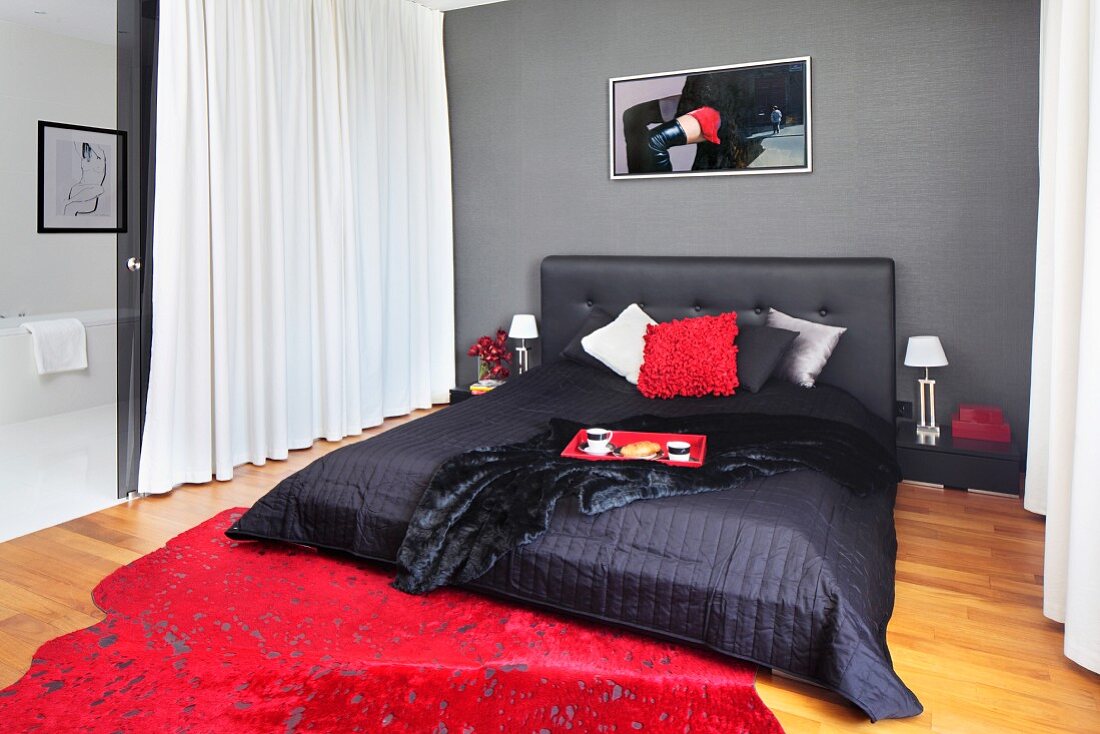 Black double bed with upholstered headboard and blanket against grey wall; red, speckled rug on floor
