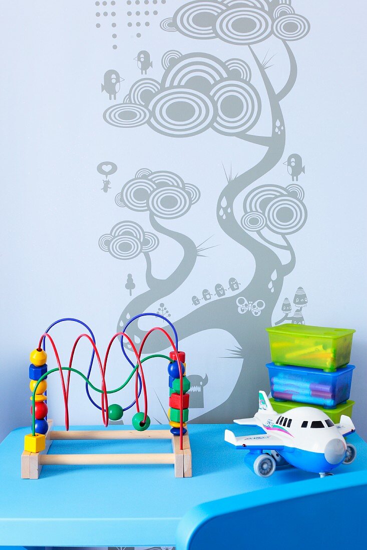 Spielzeug auf blauem Tisch, vor Wand mit Baummotiv als Schablonenmalerei