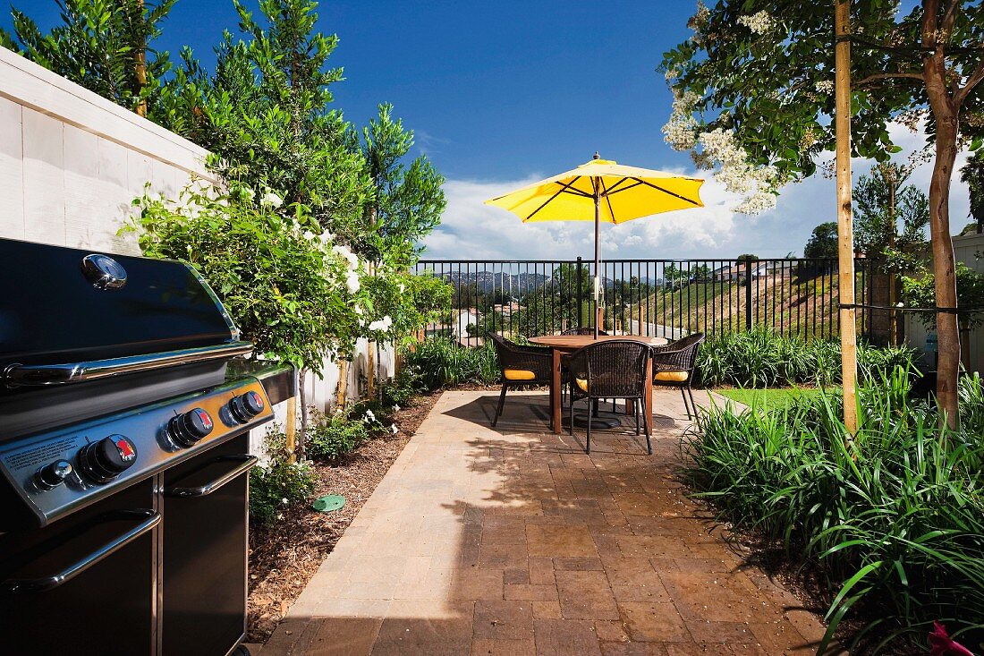 Sitzplatz mit gelbem Sonnenschirm auf sonnenbeschienener Terrasse im Garten, vorne Aussengrill an heller Steinwand