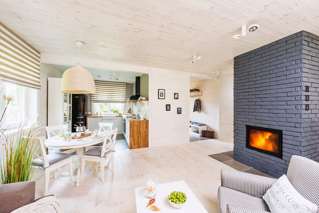 Pale, open-plan interior with fireplace built into grey brick partition and dining area below rattan pendant lamp
