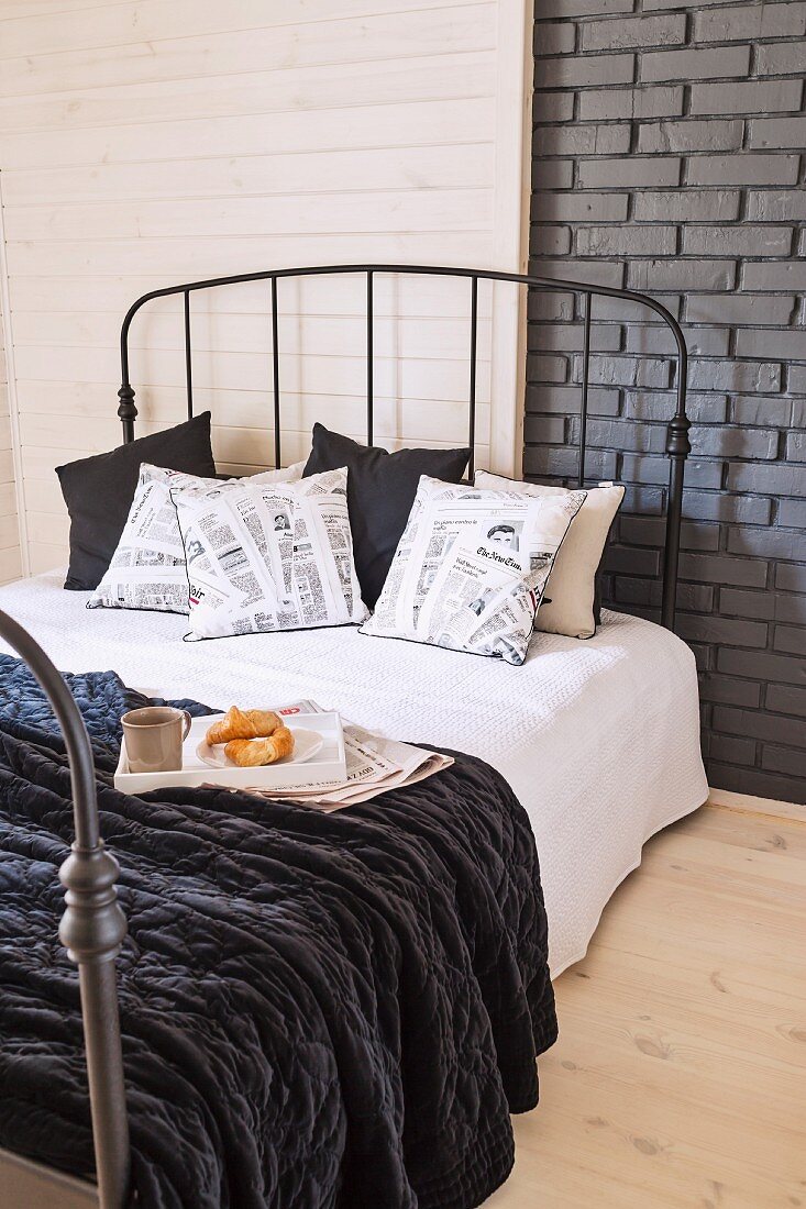 Frühstückstablett auf Bett mit dunklem Metallgestell vor Wand teilweise mit Holzverkleidung und grauer Ziegelwand