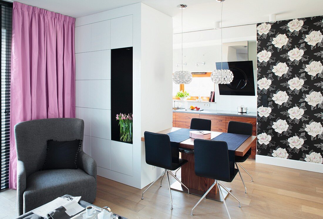 A view from a seating area looking towards a dining table with crystal pendant lamps and black and white floral wallpaper and the kitchen in the background behind a room divider cupboard